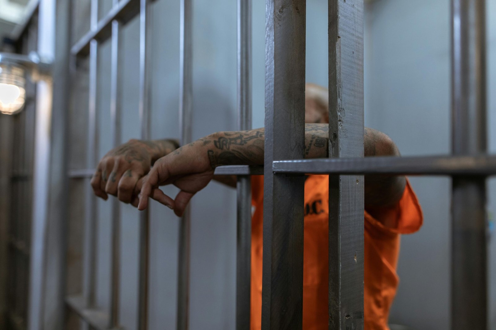 A tattooed inmate's arms rest on jail cell bars, wearing an orange uniform inside a prison.