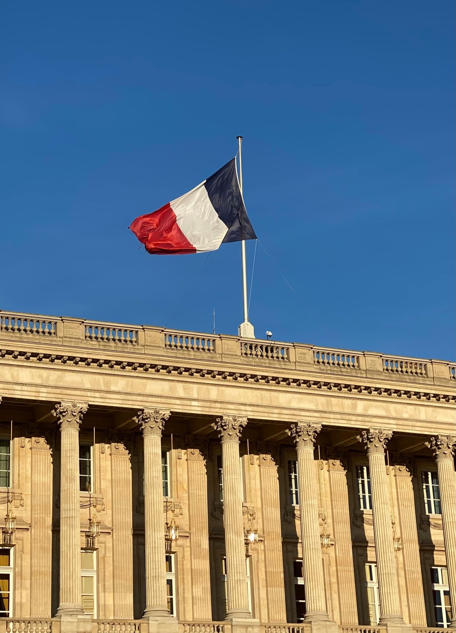 The French flag waves atop a neoclassical building against a clear blue sky.